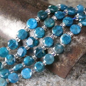 May include: Close-up of several strands of faceted, round, blue-green gemstone beads. The beads are strung together with small, clear spacer beads. The background is a textured, rusty metal surface.