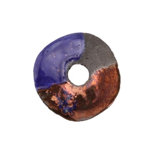 May include: A ceramic donut-shaped bead with a hole in the center. The bead is divided into three sections: a glossy blue, a matte gray, and a copper-colored section. The bead has a unique, organic shape.