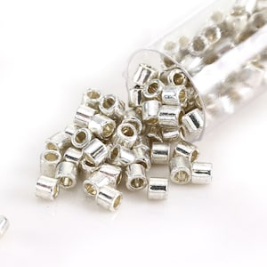 May include: A collection of small, silver-colored metal tube beads, some scattered and others spilling from a clear plastic vial. The beads have a cylindrical shape with a hole running through the center, ideal for stringing.