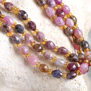 May include: Close-up of three strands of gemstone necklaces. The faceted beads are in shades of pink, purple, and brown, interspersed with small gold-colored beads. The necklaces are draped over a textured, off-white surface.