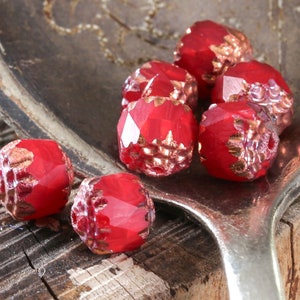 May include: A group of red glass beads with a gold metallic finish. The beads are faceted and have a textured surface. The beads are on a silver spoon.
