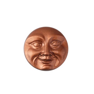May include: A bronze-colored, circular decorative element featuring a smiling face with detailed facial features. The face has a jovial expression, with visible wrinkles and a slight smirk. The item is likely a decorative accent or embellishment.