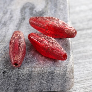 May include: Four red, oblong glass beads with a textured surface and a hole at each end. The beads have a slightly translucent appearance with a reddish-brown, speckled finish. They are arranged on a grey, textured surface.