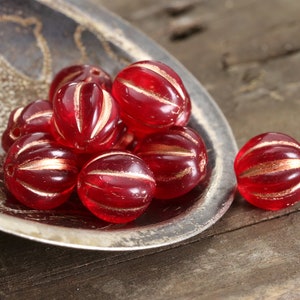 May include: A collection of red, round beads with gold stripes, displayed in a silver dish. The beads have a ribbed texture and are arranged on a rustic wooden surface. These beads are suitable for jewelry making and craft projects.