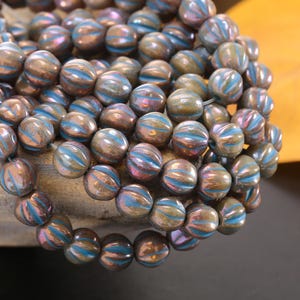 May include: A collection of round beads with a metallic finish. The beads are primarily bronze with blue and pink stripes. The beads are strung together, suggesting they are for jewelry making or crafting.