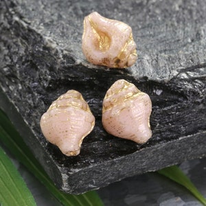 May include: Three pink seashell-shaped glass beads with gold accents. The beads are on a dark gray textured surface.
