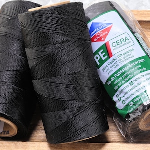 May include: Three spools of black waxed polyester thread, each labeled "Linhasita PE Cera 100% Polyester" with the text "peso liquido 100 gramas, net weight 100 grams, peso neto 100 gramos, Fio de Poliester Encerado, waxed polyester cord, hilo de poliester encerado" and contact information for Linhasita.