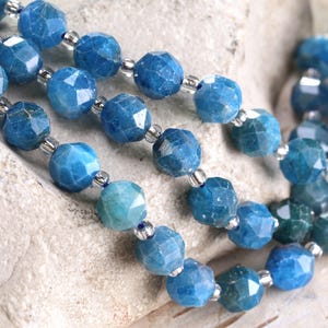 May include: Close-up of three strands of faceted blue gemstone beads. The beads are a mix of light and dark blue, with silver-colored spacers. The beads are strung on a clear, flexible cord. The background is a light-colored, textured surface.