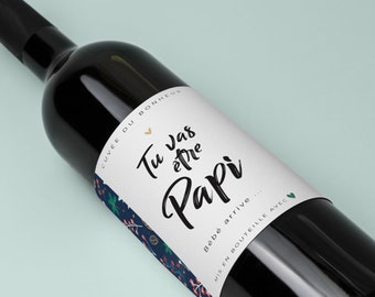 Puede incluir: Una botella de vino de vidrio oscuro con una etiqueta blanca. La etiqueta tiene el texto "Tu vas être Papi" en escritura negra, un pequeño corazón dorado y un diseño floral. La botella está sobre un fondo azul claro.