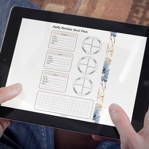 Può includere: Un tablet mostra un "Piano dei pasti giornalieri a porzioni" con sezioni per la colazione, il pranzo e la cena, insieme a una griglia e guide circolari per le porzioni di cibo. Il design include accenti floreali blu e oro.
