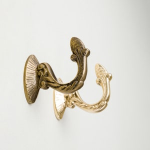 Puede incluir: Dos ganchos de metal de color dorado ornamentados con un diseño de volutas decorativas. Los ganchos están montados en una pared blanca.
