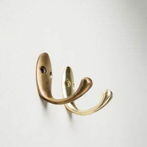 Op de afbeelding: Twee metalen haken, een gouden en een bronzen, gemonteerd op een witte muur. De haken zijn gebogen en hebben een eenvoudig ontwerp.