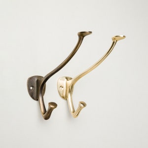 Könnte beinhalten: Zwei Metall-Kleiderhaken, einer in dunkler Bronze und der andere in glänzendem Messing. Beide haben ein gebogenes Design mit einem kleinen Haken und einer flachen Oberseite. Sie sind an einer weißen Wand montiert.