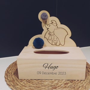 Puede incluir: Tapa de caja de pañuelos de madera con un recorte de Dumbo el elefante sosteniendo un pompón azul. La caja está personalizada con el nombre "Hugo" y la fecha "09 Décembre 2023".