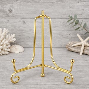 May include: A gold-colored metal display stand featuring a central vertical support, two curved side arms, and two front-facing feet with curled ends. The structure is designed to hold items upright, showcasing a smooth, reflective metallic finish.