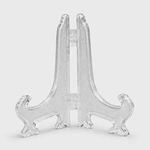 May include: Clear plastic display easel with a glittery texture. The easel has a two-piece design with a hinged back and decorative scrollwork on the feet. It is designed to hold small plates or other decorative items.