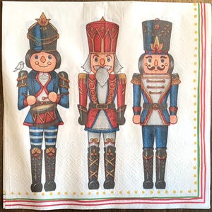 May include: Three nutcracker figurines with red, blue, and white accents. The nutcrackers are wearing traditional costumes and hats. The nutcracker on the left is holding a drum, the nutcracker in the middle is holding a sword, and the nutcracker on the right is holding a staff.