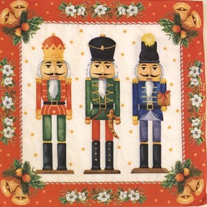 May include: A decorative Christmas napkin featuring three colorful nutcracker figures. The nutcrackers are dressed in red, green, and blue uniforms with gold accents. The napkin has a red border with bells, holly, and white flowers.