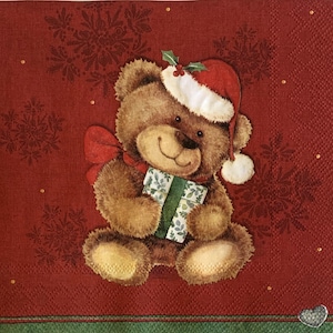 May include: A Christmas-themed napkin featuring a brown teddy bear wearing a Santa hat with holly and holding a wrapped gift. The background is red with snowflake patterns and gold accents. A red bow is around the bear's neck.