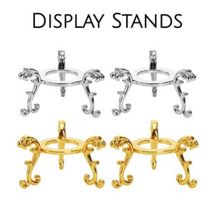 May include: Four display stands with the text DISPLAY STANDS at the top. Two stands are silver-colored and two are gold-colored. Each stand features a circular top ring supported by three ornate, curved legs with detailed scrollwork patterns.