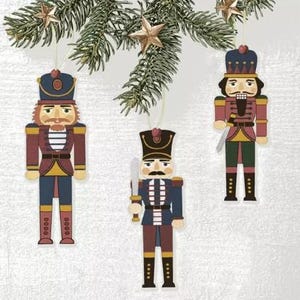 May include: Three wooden Christmas ornaments shaped like nutcracker soldiers, hanging from a pine branch. The ornaments are painted in shades of red, blue, and gold, with detailed facial features and uniform designs. Gold star ornaments are also hanging from the branch.