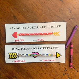 May include: Two Valentine's Day cards with the text "QUAM DULCIS ARCUS CUPIDINIS EST!" and arrow designs. One card has a red heart arrow, the other a black heart arrow. The cards are personalized with names. Two crayons are next to the cards.