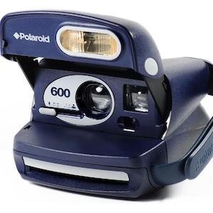 Peut inclure: Appareil photo instantané Polaroid 600 bleu marine avec flash intégré. L'appareil photo a un objectif blanc et une poignée. Le mot "Polaroid" est imprimé sur le dessus et la poignée. Le chiffre "600" est imprimé à l'avant.