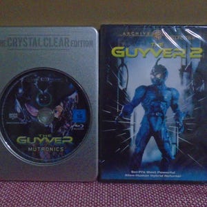 Puede incluir: Dos DVDs uno al lado del otro. El de la izquierda está en una caja plateada con el texto "THE CRYSTAL CLEAR EDITION" y "THE GUYVER MUTRONICS". El DVD de la derecha está en una caja negra con el título "THE GUYVER 2" y una imagen de un humanoide azul.