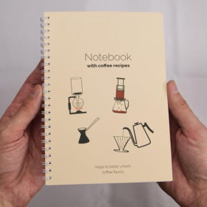 Puede incluir: Cuaderno en espiral de color crema con el texto "Notebook with coffee recipes". La portada presenta ilustraciones en negro de equipos para preparar café. El texto "Helps to better unlock coffee flavors" está en la parte inferior.
