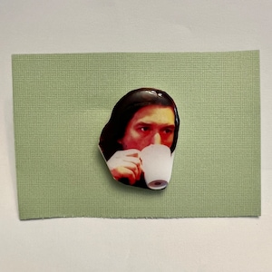 May include: A close-up of a man with dark hair drinking from a white mug. He is wearing a dark shirt and has a serious expression on his face.