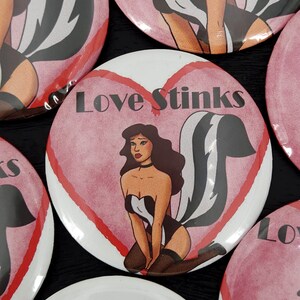May include: A pink and white heart-shaped button with a black and white skunk illustration and the text "Love Stinks". The skunk is wearing a white and black dress and has a pink heart on its tail.