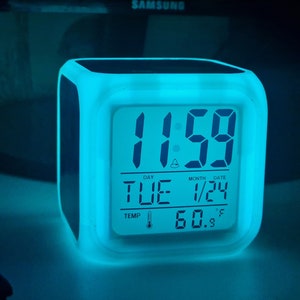 Puede incluir: Un reloj despertador digital blanco con retroiluminación azul muestra la hora 11:59, el día "TUE", el mes y la fecha "1/24", y la temperatura "60.9°F".
