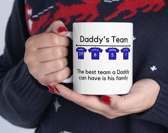 Op de afbeelding: Een witte keramische mok met een blauw-wit ontwerp. De mok heeft de tekst "Daddy's Team" en een rij van vier blauwe shirts met namen en nummers erop. De tekst onder de shirts luidt "Het beste team dat een vader kan hebben is zijn familie."