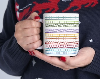 Puede incluir: Taza de cerámica blanca con un patrón de líneas horizontales de colores y puntadas decorativas. La taza tiene un asa blanca. La taza es sostenida por una persona que lleva un suéter azul oscuro.