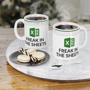 Puede incluir: Dos tazas de café blancas con un logotipo de Excel verde y el texto "Freak in the sheets". Las tazas están sobre una superficie de mármol con un plato de macarons y una cuchara.