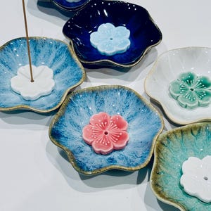 May include: A collection of six ceramic incense holders shaped like flower petals. Each holder features a central raised flower-shaped piece with a small hole for an incense stick. The dishes come in various glazed finishes including blue, white, and green.