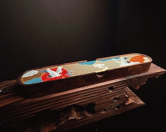 Puede incluir: Una bandeja de té de madera de forma ovalada y alargada con una superficie de listones. La bandeja está decorada con un estampado gráfico colorido de grullas blancas, un gran sol rojo y nubes azules estilizadas sobre un fondo de madera natural.