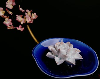 May include: A ceramic incense holder featuring a deep blue, leaf-shaped dish with a white lotus flower sculpture in the center. A thin, gold-toned stem extends from the dish, topped with a cluster of small, delicate pink and white blossoms.