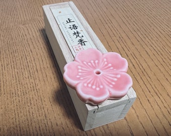May include: A light pink ceramic flower incense holder with a central hole, resting on a rectangular light-coloured wooden box. The box features a white label with black Chinese characters and the text RHYME SANSKIRT printed in black ink.