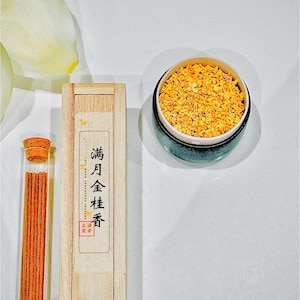 May include: A still-life image featuring incense and related items. A wooden box with Chinese characters and a glass tube with incense sticks are next to a small ceramic container filled with small, orange-coloured pieces.