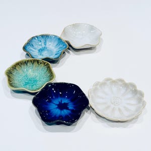 May include: Six small ceramic dishes in various colours, including blue, white, and green. Each dish has a unique shape, with a scalloped or floral design. The dishes are arranged on a white surface, highlighting their individual patterns and textures.