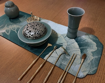 Puede incluir: Un quemador de incienso de cerámica verde con una tapa de metal y un diseño floral, un pequeño frasco de cerámica verde, una taza de cerámica verde y seis herramientas de incienso de metal dorado sobre una superficie con estampado verde y dorado.