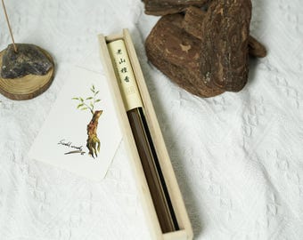 May include: A rectangular light-coloured wooden box containing dark incense sticks, accompanied by a white card featuring a botanical illustration and the text "Sandal wood," alongside a small incense holder and wood pieces.