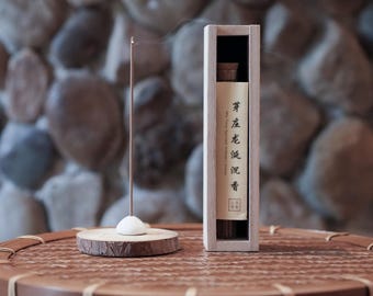 Può includere: Un sottile bastoncino di incenso brucia in un supporto bianco a forma di cupola posto su una base circolare in legno. Accanto si trova una scatola alta e rettangolare in legno chiaro contenente incenso, con un'etichetta di carta con testo giapponese.