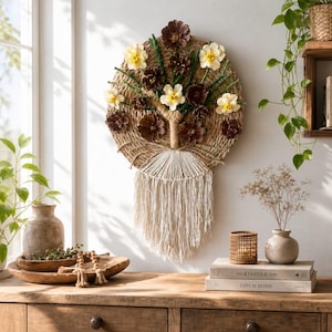 May include: A circular wall hanging featuring a woven jute base decorated with dark brown pinecones and cream-colored fabric flowers on green stems. The lower section consists of white macrame fringe hanging from a central wrapped jute trunk.