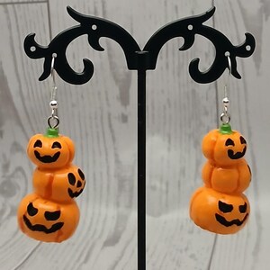 May include: A pair of dangle earrings featuring a vertical stack of three orange jack-o'-lantern pumpkins with green stems and black painted facial expressions. The charms are suspended from silver-toned metal fishhook ear wires.