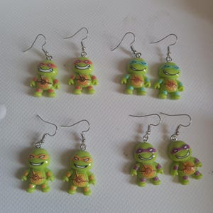 Puede incluir: Seis pares de pendientes de las Tortugas Ninja. Cada pendiente presenta una pequeña figura de tortuga sonriente con una máscara de color y un gancho plateado. Las tortugas son verdes y amarillas con máscaras rojas, azules, naranjas y moradas.