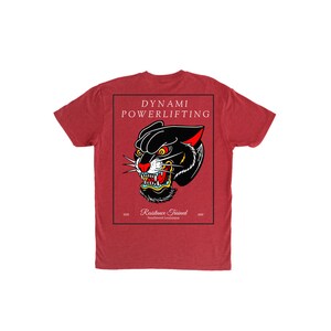 Könnte beinhalten: Rotes T-Shirt mit einem schwarzen Pantherkopf-Motiv, gelben Augen, roten Akzenten und scharfen Zähnen. Das Shirt zeigt den Text "DYNAMi POWERLIFTING" und "Resistance Trained Southwest Louisiana".