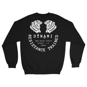 Könnte beinhalten: Schwarzes Sweatshirt mit einer weißen Grafik, die zwei brüllende Löwenköpfe und den Text "DYNAMi RESISTANCE TRAINED" in einem halbkreisförmigen Design zeigt. Das Sweatshirt enthält auch die Adresse "000 BROAD STREET STHWST - LA 70600".