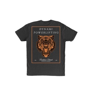 Könnte beinhalten: Dunkelgraues T-Shirt mit einem orangefarbenen Tigerkopf-Motiv in einem rechteckigen Rahmen. Der Text "Dynami Powerlifting" steht über dem Tiger, darunter "Resistance Trained Southwest Louisiana". Das T-Shirt hat einen Rundhalsausschnitt.